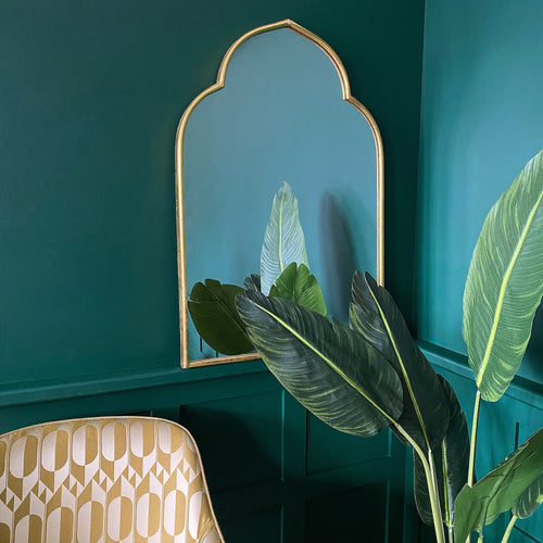 A gold Moroccan arch mirror on the wall with a chair and plants in front of it