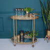 A gold bar cart with two shelves, holding decorative glasses and plant pots