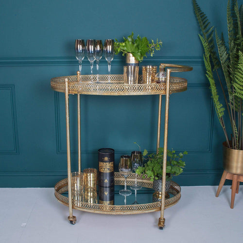A gold bar cart with two shelves, holding decorative glasses and plant pots
