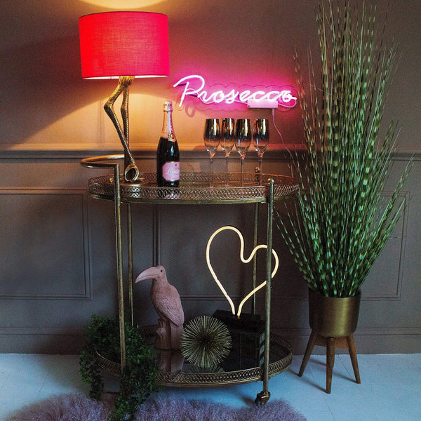A gold bar cart with various decorative items including wine glasses, a table lamp, and a neon sign behind it