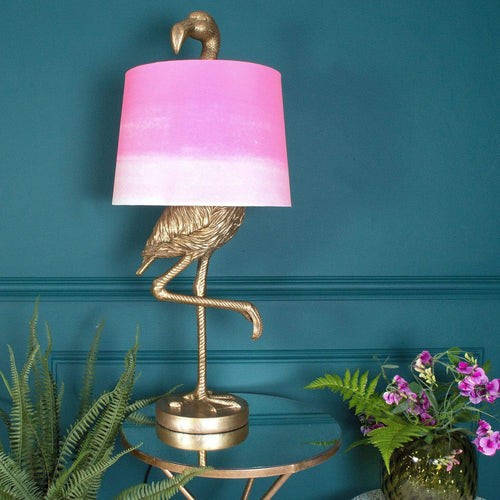 A gold flamingo lamp with pink shade sits on a table beside a vase
