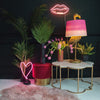 A gold flamingo lamp with a pink shade standing on a table beside plants and neon lights
