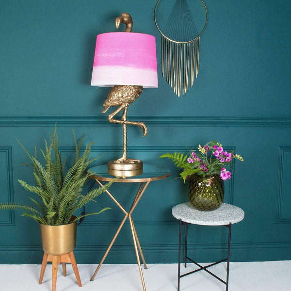 A gold flamingo lamp with a pink shade on a table beside plants