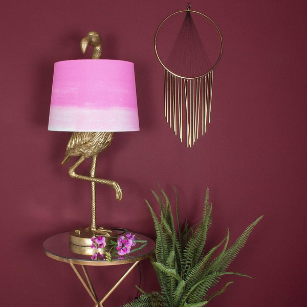 A gold flamingo lamp with a pink shade next to a gold wall decor and plants