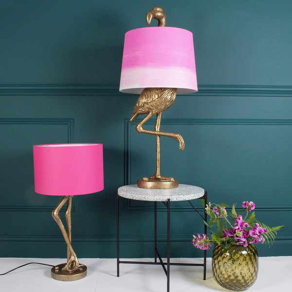 A gold flamingo lamp with a pink shade on a table next to another lamp and a vase