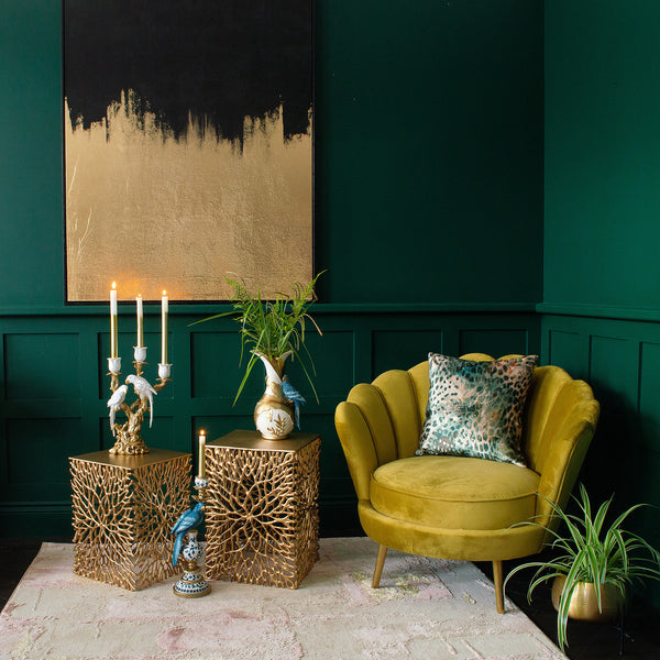 A yellow chair, a cushion, side tables, candle holders, plant pots, and a gilded canvas art on the wall