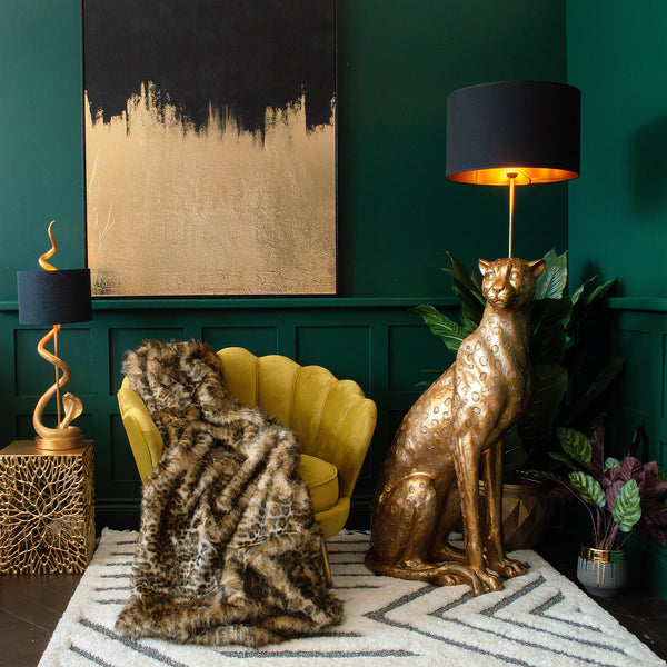 A yellow chair, a cheetah floor lamp, a table lamp on a side table, plant pots, and a gilded canvas art