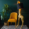 A golden giraffe floor lamp with a black shade next to an orange chair and a plant pot