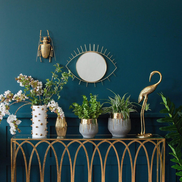A console table with various plant pots and vases on it, and a mirror and gold beetle wall decor on the wall