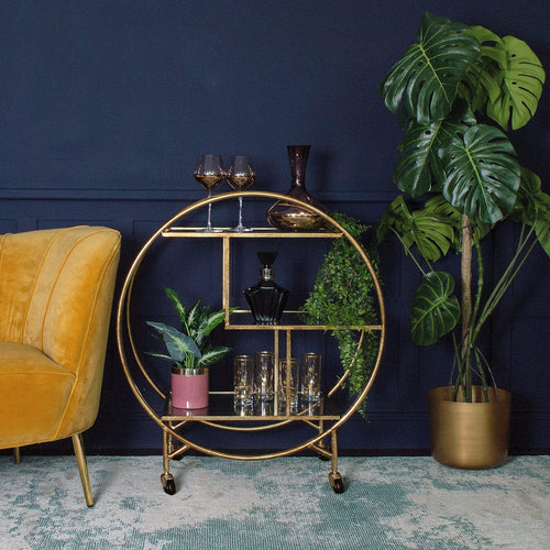 A stylish gold round drinks trolley next to a plant pot and a chair