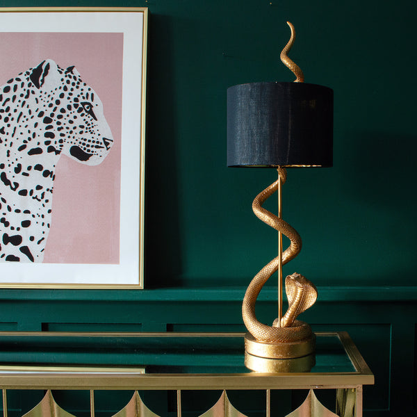 A gold snake table lamp with a black shade on a console table, and a leopard print on the wall