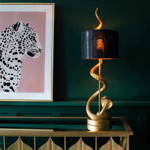 A gold snake table lamp with a black shade on a console table, and a leopard print on the wall