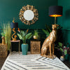A room with plants, a round wall mirror, a cheetah-shaped lamp, a gold snake table lamp, and a rug