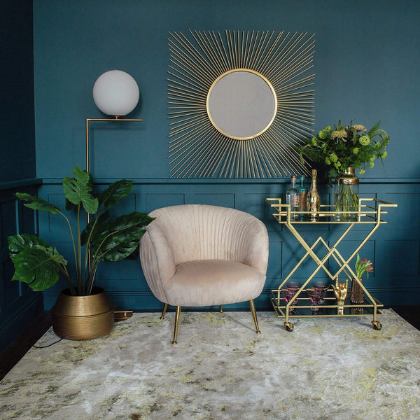 A room with an armchair, wall mirror, floor lamp, plant pot, a gold bar cart, and a golden lustre rug