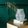 A glass bum vase with plants inside it is placed on a stack of books on a table