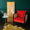 A red armchair is placed next to a golden framed artwork and a small white side table with a glass bum vase on it