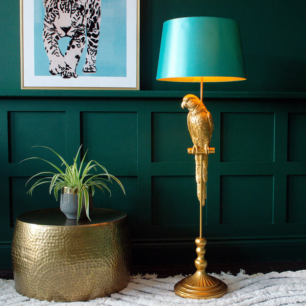 A golden parrot floor lamp with a turquoise shade next to a plant pot and a framed leopard print