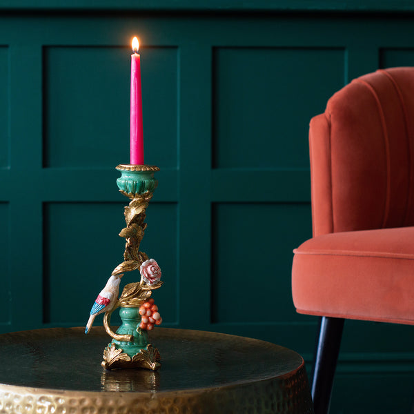 A perching bird candle holder with a pink candle on a table next to a red chair