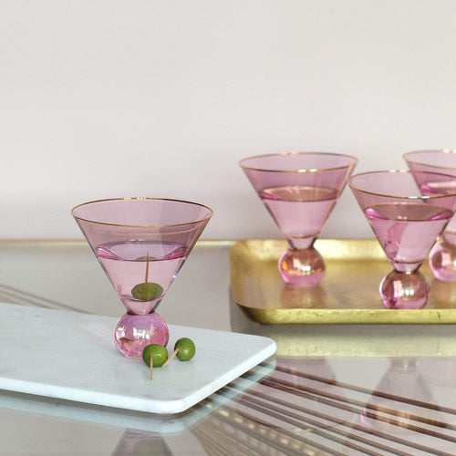 Four pink martini glasses with gold rims, one on a marble slab with an olive skewer, and three on a gold tray