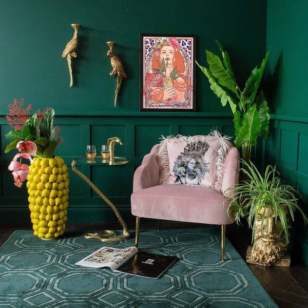 A room featuring a pink chair, a side table, a lemon yellow vase, wall decor items, and plants