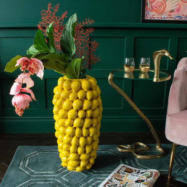 A lemon yellow vase with flowers inside it next to a side table and a chair