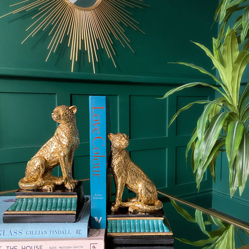 Two gold leopard bookends hold up a book on a glass table and a large plant beside it