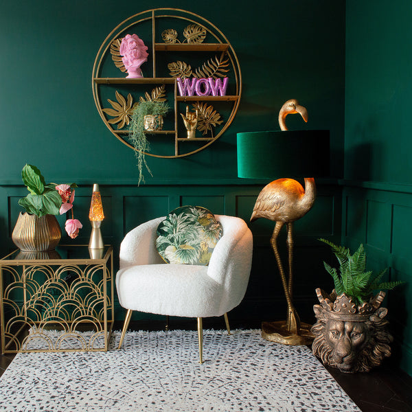 A room featuring a white armchair, gold side table, flamingo lamp, a wall shelf, and eclectic decor